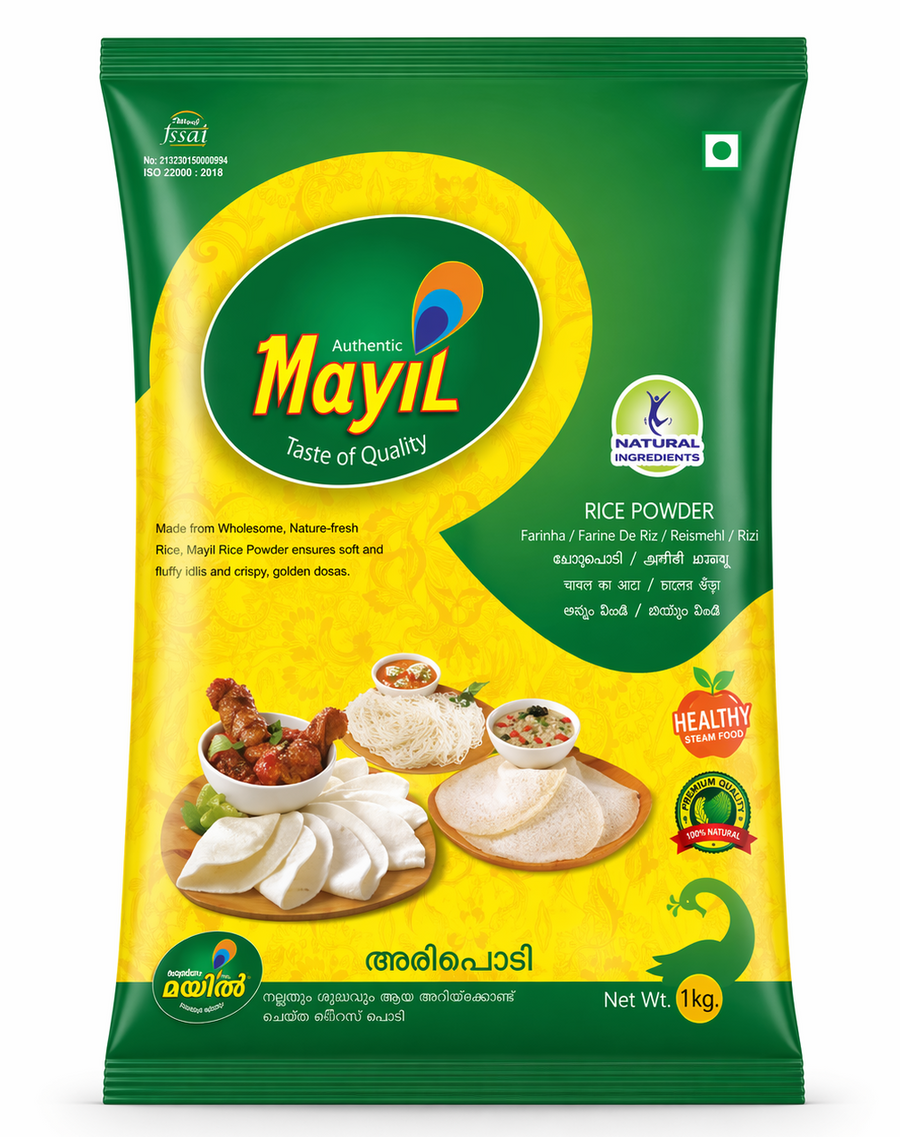 A high-quality studio shot of a Mayil Rice Powder. The packaging features a vibrant green and yellow design with a large green oval containing the "Mayil" peacock logo. The center image showcases a variety of prepared South Indian dishes, including soft white Idlis, lacy Appams, and delicate Idiyappams, accompanied by bowls of sambar and chutney. Several quality seals, such as "Natural Ingredients," and "100% Natural," are prominently displayed. This product is available in the UK at theindianshelf.co.uk.