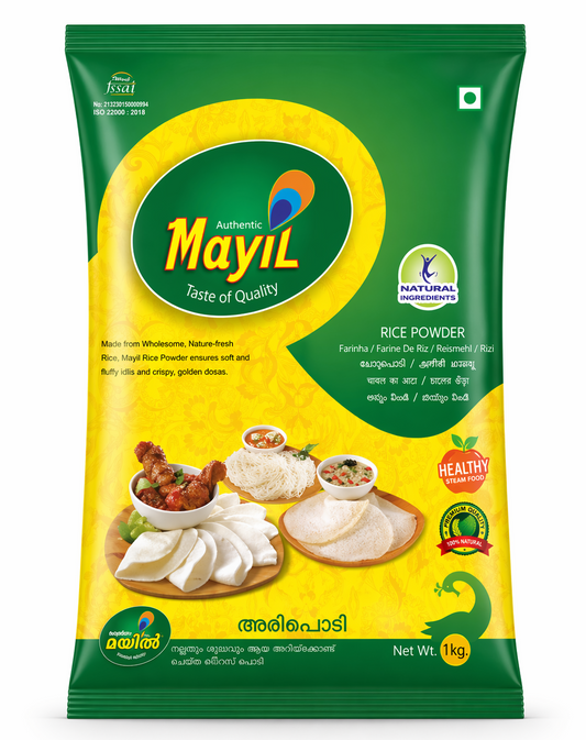A high-quality studio shot of a Mayil Rice Powder. The packaging features a vibrant green and yellow design with a large green oval containing the "Mayil" peacock logo. The center image showcases a variety of prepared South Indian dishes, including soft white Idlis, lacy Appams, and delicate Idiyappams, accompanied by bowls of sambar and chutney. Several quality seals, such as "Natural Ingredients," and "100% Natural," are prominently displayed. This product is available in the UK at theindianshelf.co.uk.