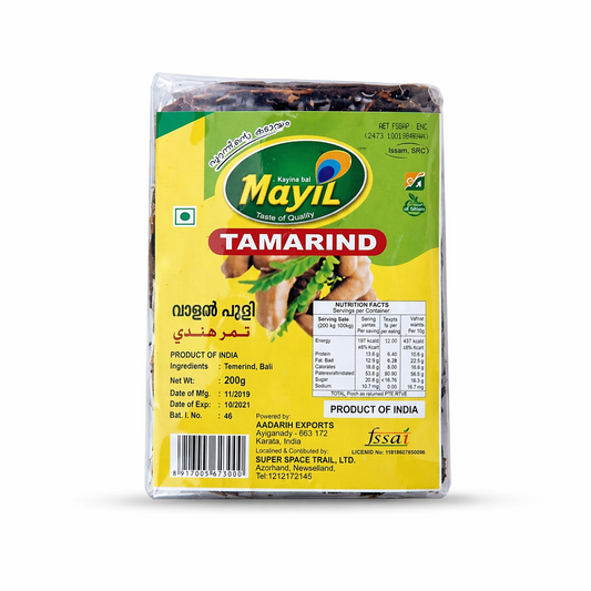 A studio shot of a Mayil Tamarind packet. The clear plastic packaging reveals dark, compressed tamarind pulp inside. The front label is bright yellow and green, featuring the "Mayil" logo with a peacock feather. Large red text reads "TAMARIND," with the name also written in Malayalam and Arabic. The label includes a nutrition facts table and confirms the net weight as 200g. This product is available in the UK at theindianshelf.co.uk.