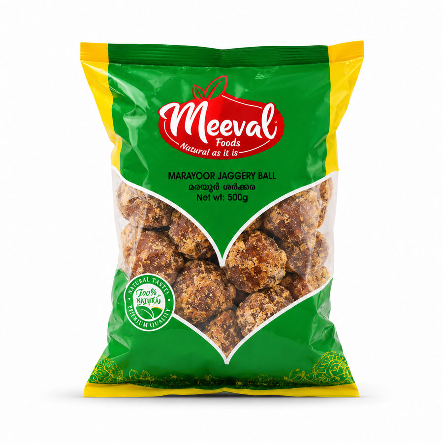 A professional studio product shot of a 500g pouch of Meeval Foods Marayoor Jaggery Ball isolated against a pure white background in image_81efe0.jpg. The green, yellow, and red packaging includes a large, clear V-shaped window showing the rustic brown jaggery balls inside.