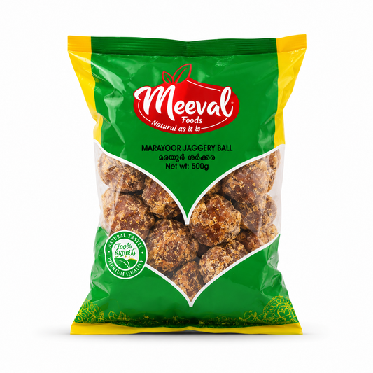 A professional studio product shot of a 500g pouch of Meeval Foods Marayoor Jaggery Ball isolated against a pure white background in image_81efe0.jpg. The green, yellow, and red packaging includes a large, clear V-shaped window showing the rustic brown jaggery balls inside.