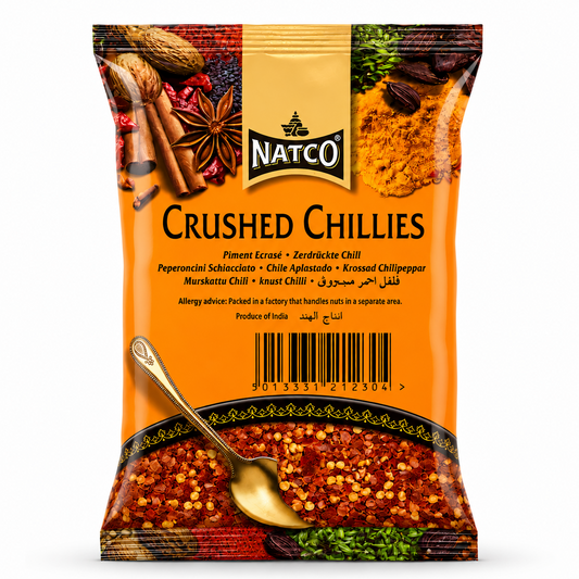 A professional studio shot of a Natco Crushed Chillies spice. The packaging features a vibrant orange background with a decorative top border showcasing various whole spices like cinnamon, star anise, and turmeric. The "NATCO" logo is prominently displayed in the center. A clear window at the bottom, styled like a bowl with a golden spoon, reveals the bright red crushed chilli flakes and seeds inside. This product is available in the UK at theindianshelf.co.uk.