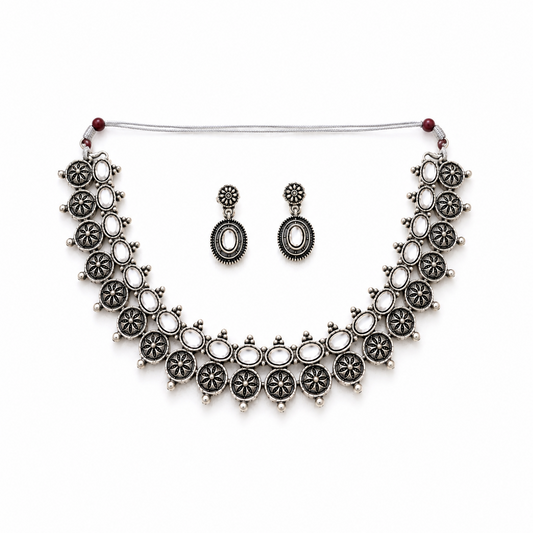 A studio product shot of an antique oxidized silver Choker Necklace and Earrings Set isolated on a white background. The jewelry features sculpted floral metalwork details set with clear white stones. Available in the UK at theindianshelf.co.uk.