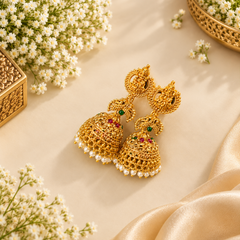 A pair of traditional matte gold-finish temple jhumka earrings featuring goddess and peacock motifs with ruby, emerald, and pearl fringe accents, displayed on a neutral background. Available in the UK at theindianshelf.co.uk.