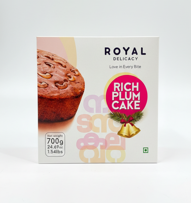 A square white box of Royal Delicacy Rich Plum Cake, set against a plain white background. The front features a close-up photograph of a section of a plum cake topped with cashew nuts. A pink circular label with a Christmas bell graphic identifies the product as "RICH PLUM CAKE." The net weight is specified as 700g (24.69 oz, 1.54 lbs). Large, faint Malayalam script is visible in the center.