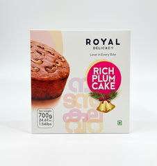 A square white box of Royal Delicacy Rich Plum Cake, set against a plain white background. The front features a close-up photograph of a section of a plum cake topped with cashew nuts. A pink circular label with a Christmas bell graphic identifies the product as "RICH PLUM CAKE." The net weight is specified as 700g (24.69 oz, 1.54 lbs). Large, faint Malayalam script is visible in the center.