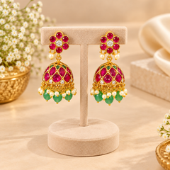 A pair of gold-plated South Indian temple jhumka earrings with ruby stones, emerald beads, and pearl drops on a beige T-bar stand. Available in the UK at theindianshelf.co.uk.
