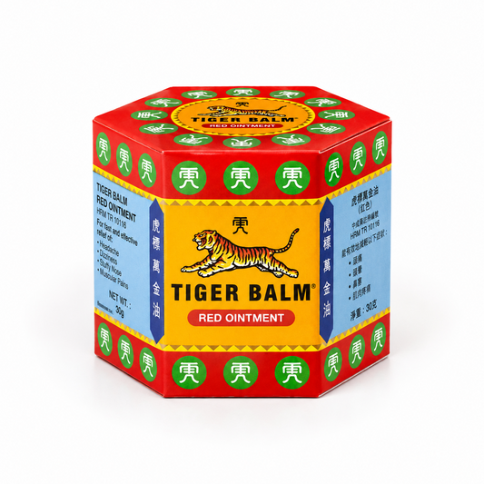 A professional studio shot showcasing the iconic, hexagonal cardboard outer carton for Tiger Balm Red Ointment The box features a striking geometric layout with bright red borders on the top, bottom, and edges, populated with circular green emblems housing white Chinese characters.The main front-facing panel has a warm golden-yellow background, framed by dark blue vertical columns containing gold Chinese script. This product is available in the UK at theindianshelf.co.uk.