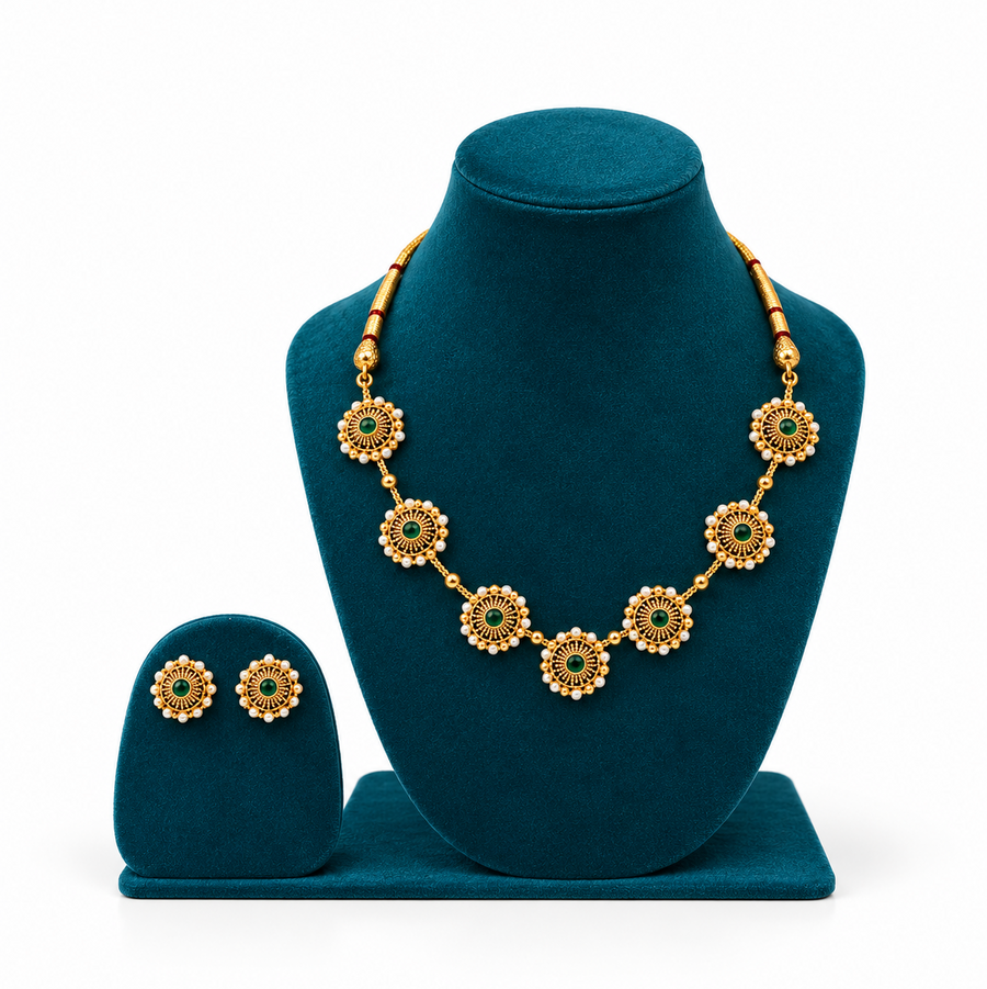 An antique gold-plated necklace set featuring circular sunburst medallions with emerald green centers and pearl borders on a peacock-blue velvet bust. Available in the UK at theindianshelf.co.uk.