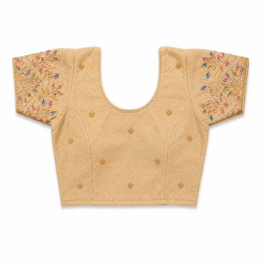 A beige short-sleeved stretchable readymade saree blouse with hand-embroidered golden leaf patterns on the sleeves and small gold floral motifs across the front, laid flat against a solid white background. Available in the UK at theindianshelf.co.uk.