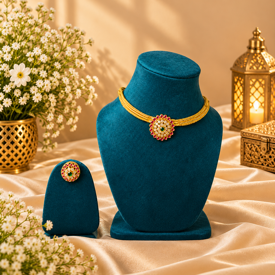 A gold-finish traditional necklace and matching earring set featuring a circular floral pendant adorned with ruby, emerald, and white stones, displayed on deep teal velvet stands. Available in the UK at theindianshelf.co.uk.