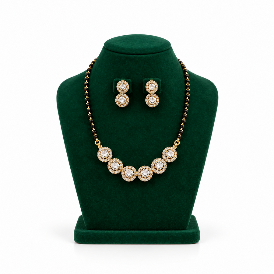 A gold-plated mangalsutra necklace with black beads and a row of circular CZ stone medallions displayed on a green velvet bust with matching earrings. Available in the UK at theindianshelf.co.uk.