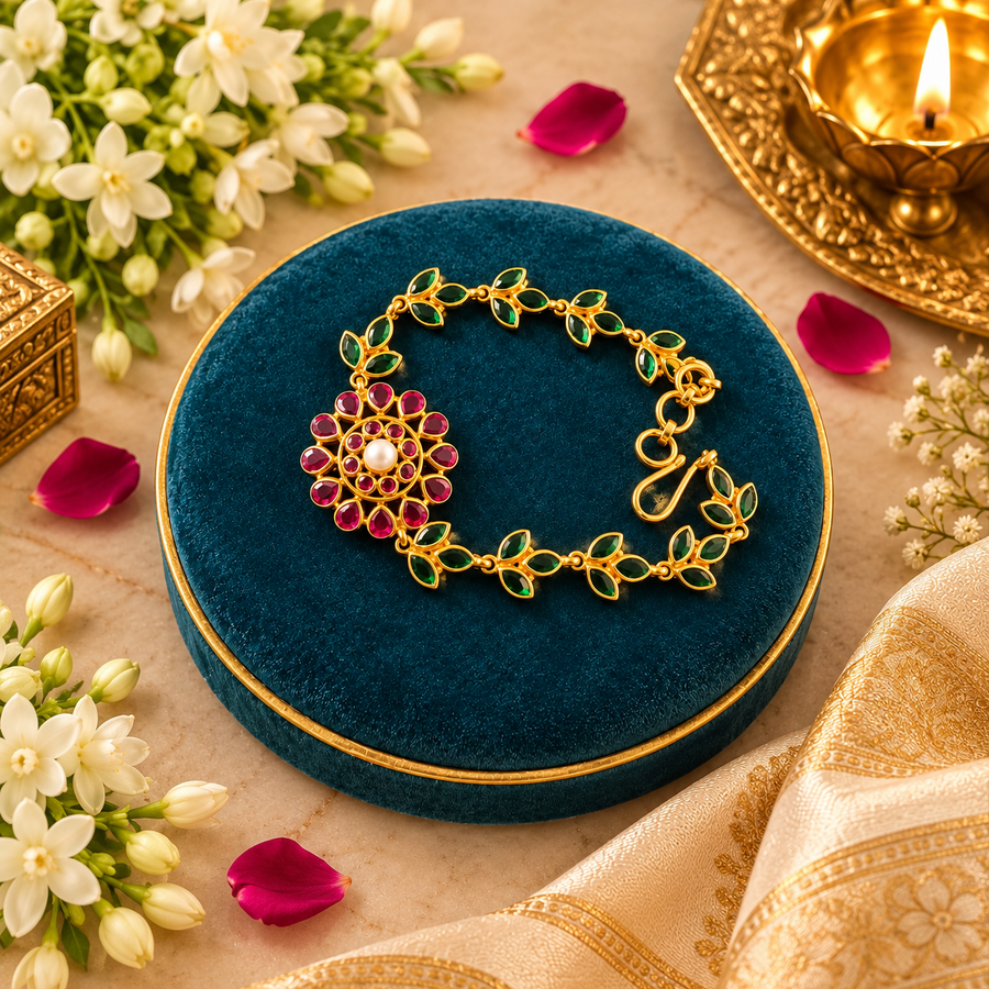 A gold-tone ethnic bracelet featuring a ruby-and-pearl floral centerpiece and green leaf links, displayed on a teal velvet surface with floral and candlelight accents. Available in the UK at theindianshelf.co.uk.