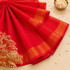A studio flatlay shot of a neatly folded crimson red Kerala Cotton Saree with subtle gold dots. The fabric features a prominent horizontal border with fine parallel gold zari stripes. A gold dish with white pearls and dried flowers decorate the background. Available in the UK at theindianshelf.co.uk.
