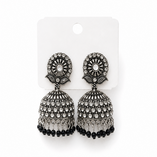 A studio product shot of a pair of antique silver oxidized Rossy Jhumka Earrings on a white display card. The dome earrings feature rows of white crystals and a fringe of small black beads. Available in the UK at theindianshelf.co.uk.