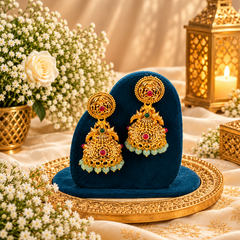 A pair of traditional matte gold-tone jhumka earrings featuring intricate filigree work, ruby and emerald stones, and soft green bead fringes, displayed on a deep teal velvet stand. Available in the UK at theindianshelf.co.uk.