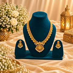 A traditional matte gold-tone long coin haram necklace and matching pearl-fringed drop earrings displayed on deep teal velvet stands. Available in the UK at theindianshelf.co.uk.
